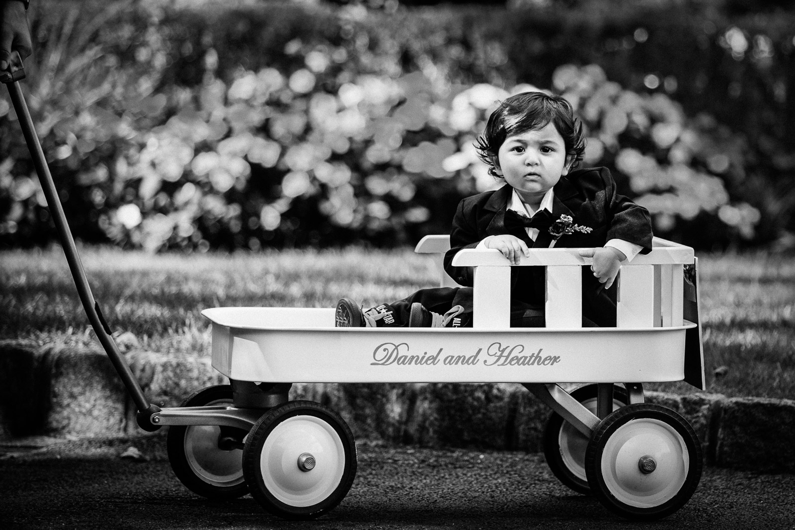 Ring bearer toddler in tuxedo sitting in personalized wagon at Crystal Plaza wedding in Livingston NJ, captured in black and white