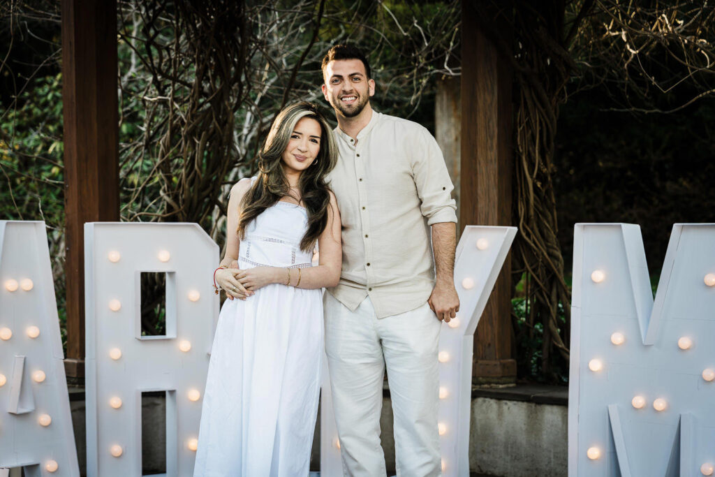 Newly engaged couple smiling in front of illuminated marquee letters at Van Vleck House Gardens
