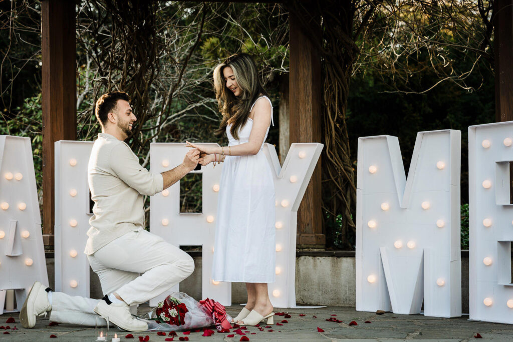 Man kneeling and placing engagement ring on partner's finger at Van Vleck House Gardens NJ