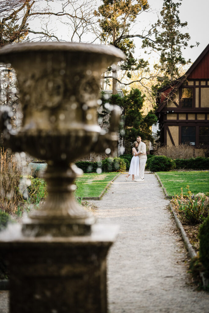 Engaged couple embracing on garden path framed by stone urn at Van Vleck House Gardens NJ