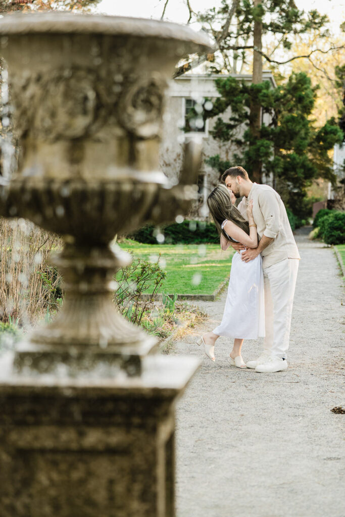 Couple kissing on garden path with ornate stone urn in foreground at Van Vleck House Gardens NJ