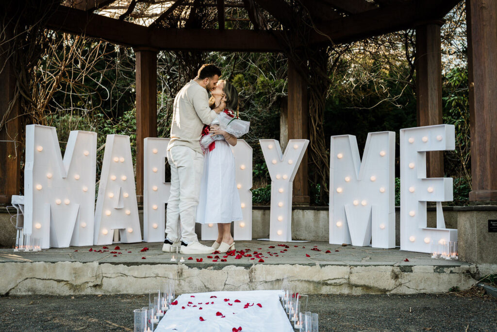 Couple sharing first kiss after surprise proposal at Van Vleck House Gardens marquee letters NJ