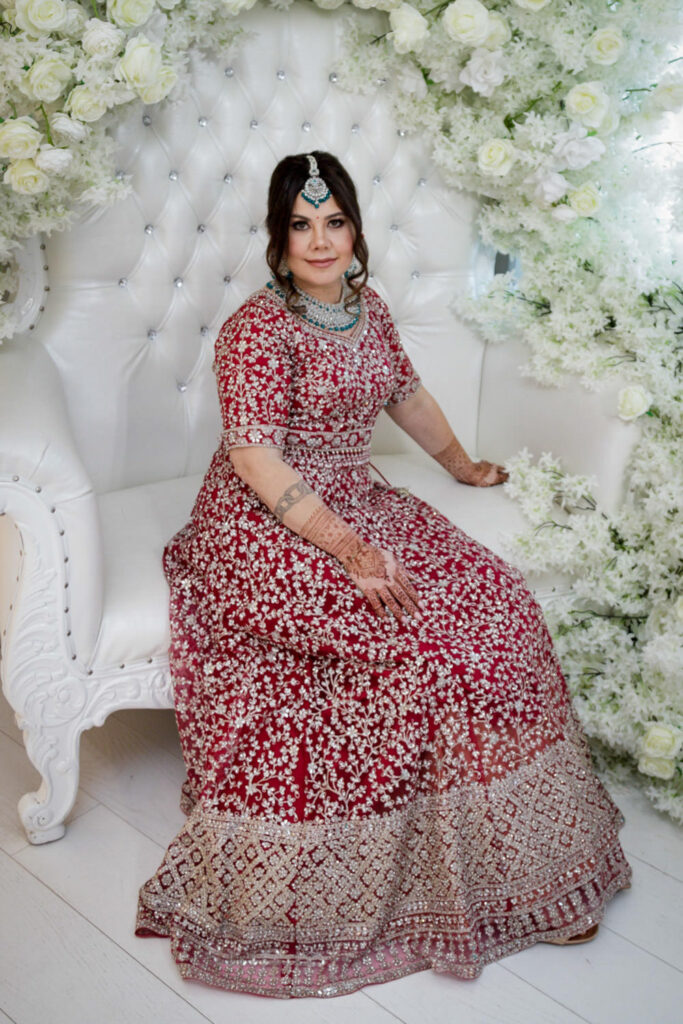 Indian bride seated in elaborate red lehenga with teal maang tikka surrounded by white floral backdrop at Crystal Plaza