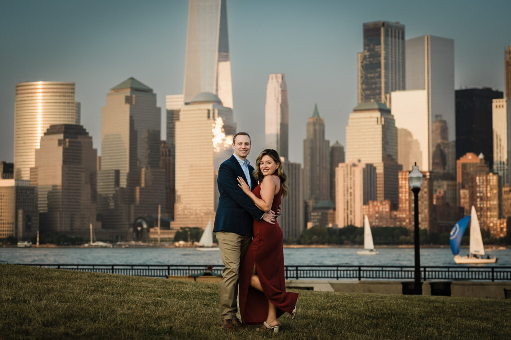 Couple embracing on the grass at Liberty State Park with sailboats and NYC skyline during engagement photos