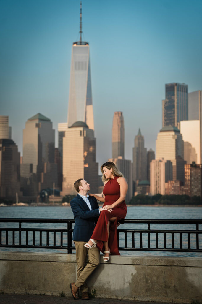 Couple at the Liberty State Park waterfront with Ivonne sitting on the ledge and NYC skyline behind them