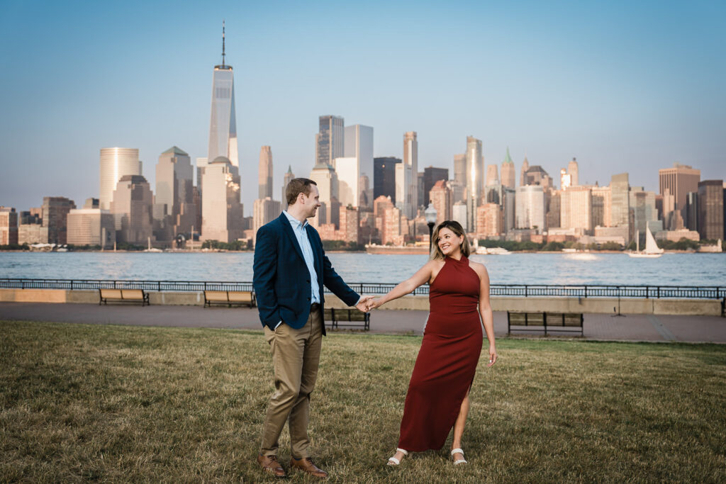 Couple holding hands on the grass at Liberty State Park with the full NYC skyline and water behind them