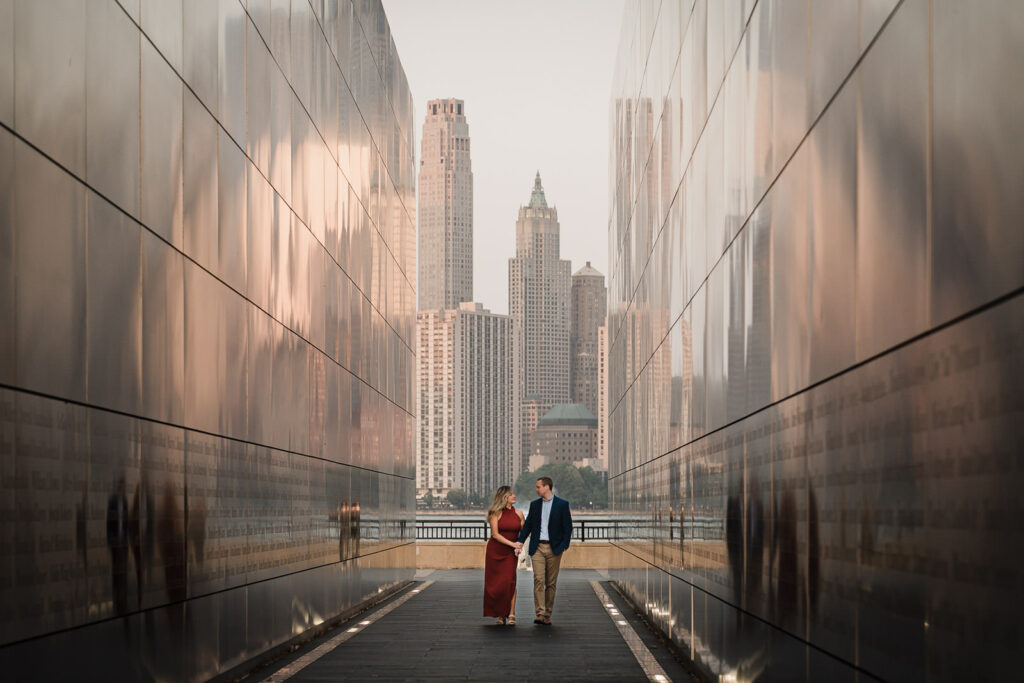 Wide shot of couple walking through the Empty Sky Memorial corridor with NYC skyline visible at the end