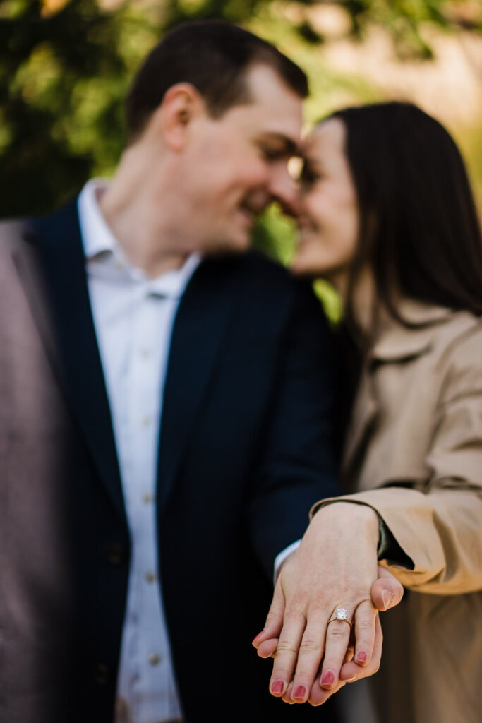 Close-up of engagement ring in sharp focus with smiling couple softly blurred in background at The Cloisters