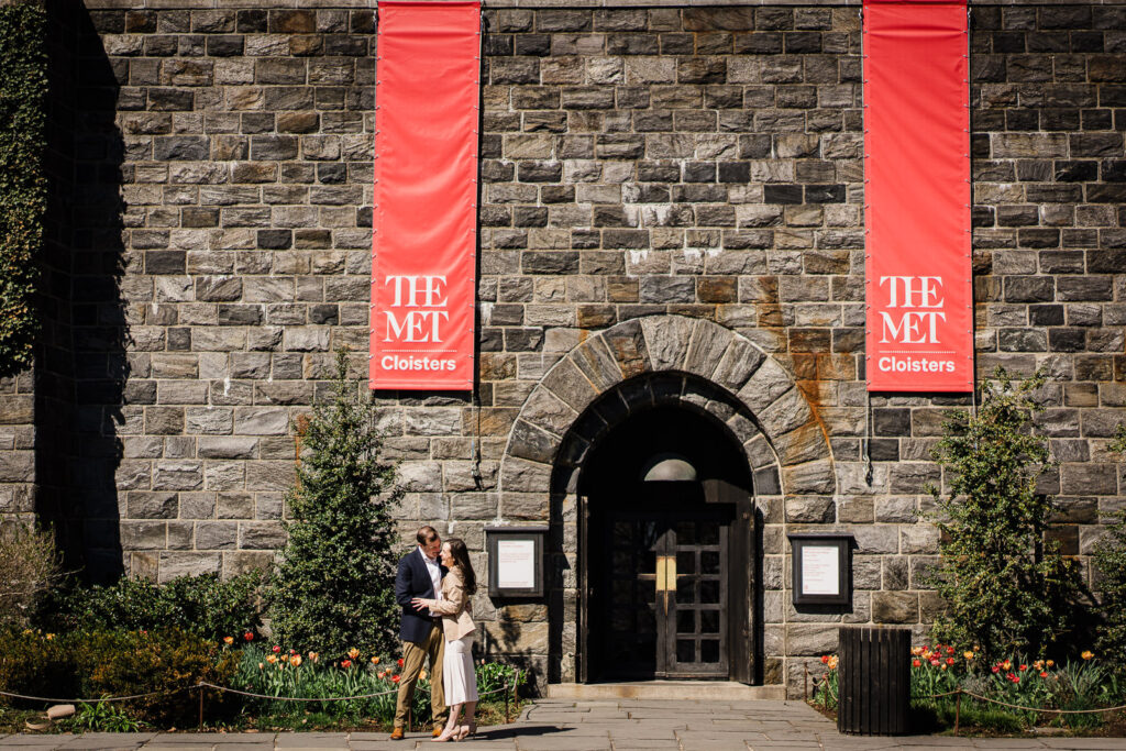 Engaged couple embracing at The Met Cloisters stone entrance beneath large red museum banners