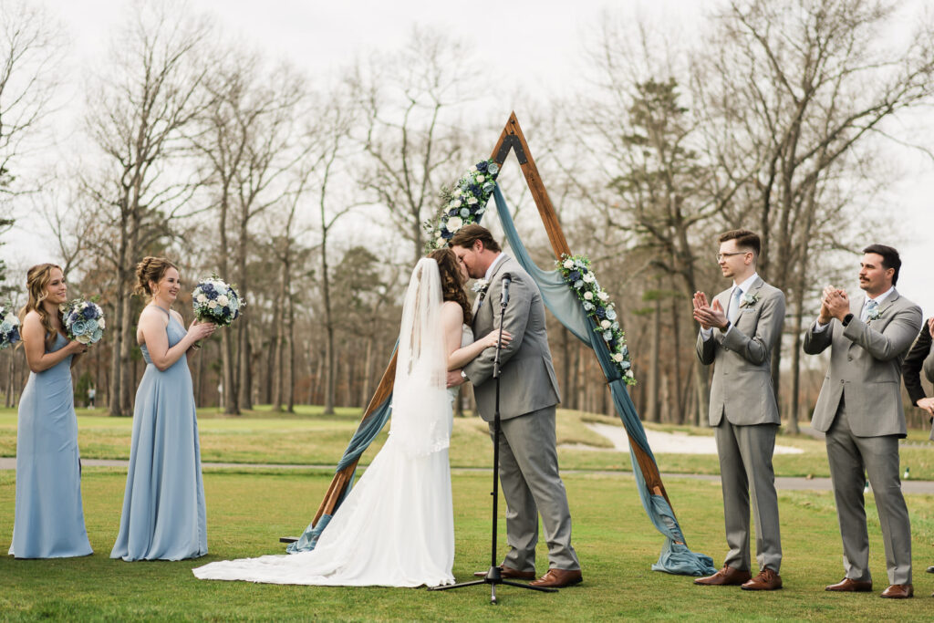 Bride and groom first kiss under triangle arch outdoor ceremony at Pinelands Golf Club NJ with wedding party