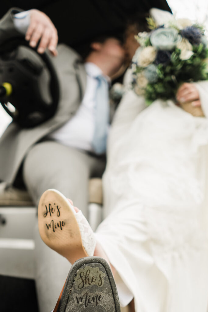 Bride shoe sole reading He's Mine with couple kissing in background inside golf cart at Pinelands Golf Club NJ