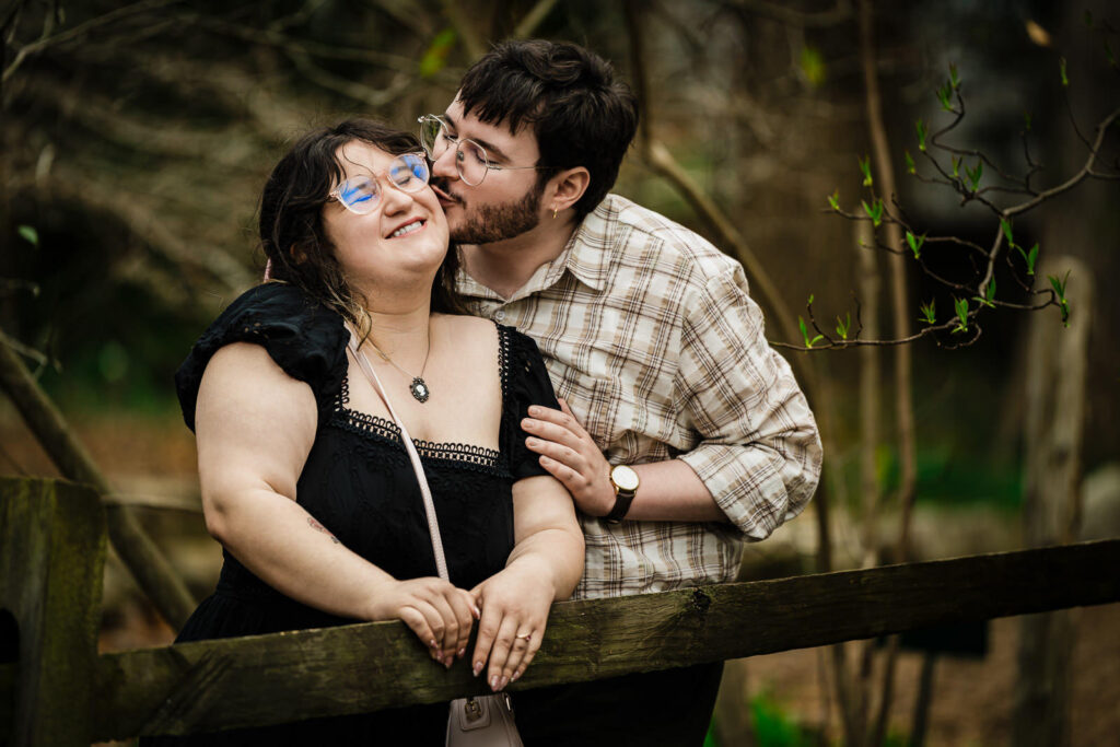 Man kissing partner on cheek while she smiles at rustic wooden fence in Reeves-Reed Arboretum NJ