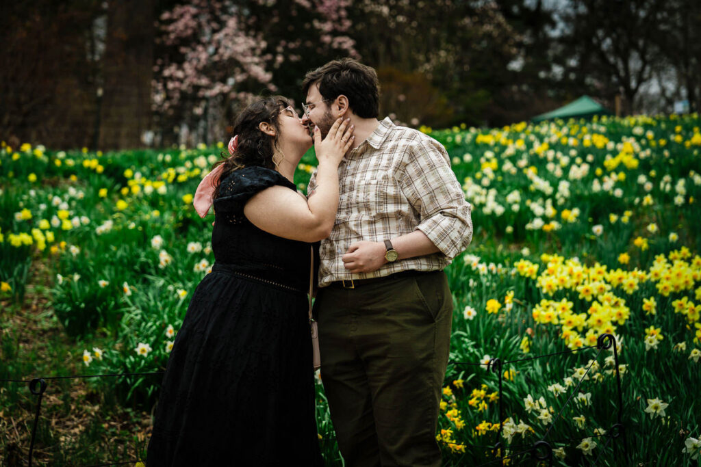 Couple sharing romantic kiss in front of daffodil garden with pink magnolia tree in background NJ