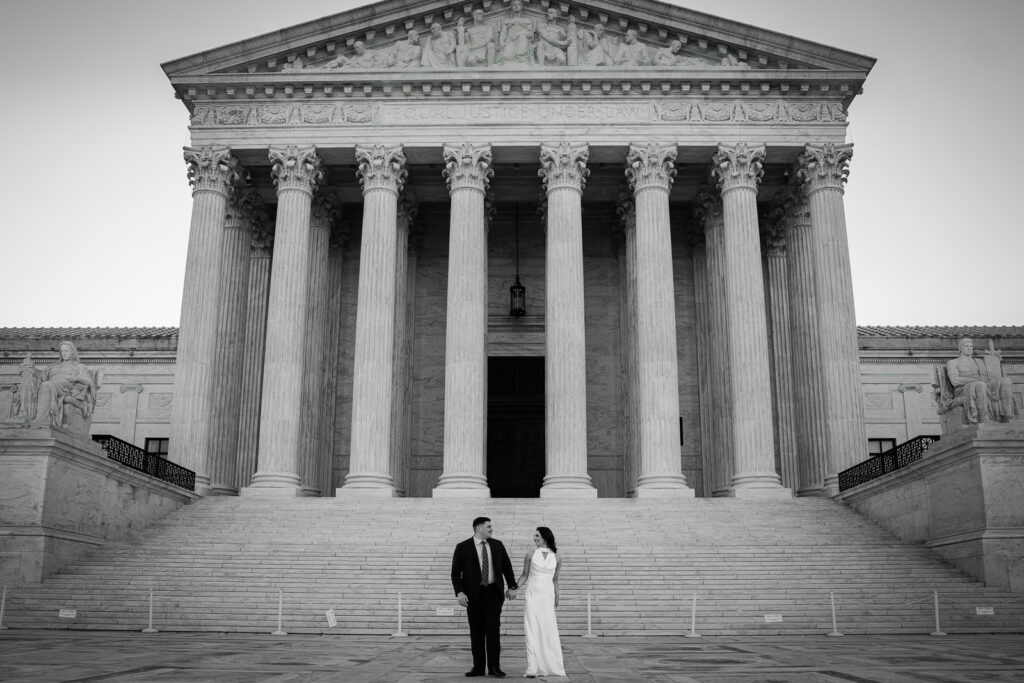 Black and white wide shot of couple holding hands facing each other in front of the full Supreme Court facade