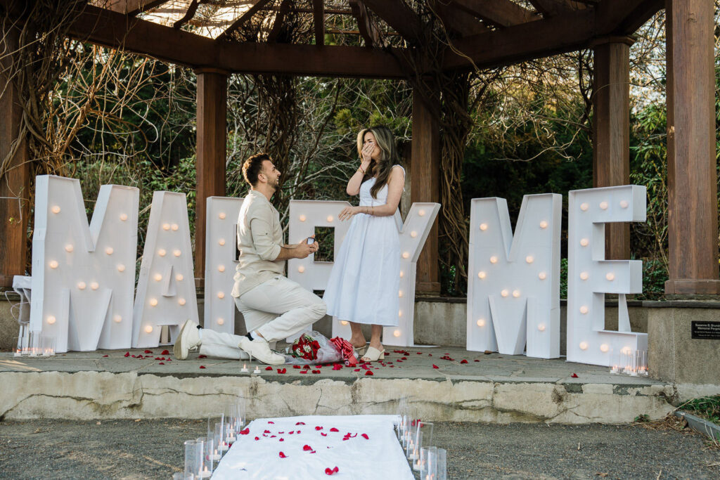 Man kneeling with ring box as woman covers mouth in surprise at Van Vleck House Gardens NJ