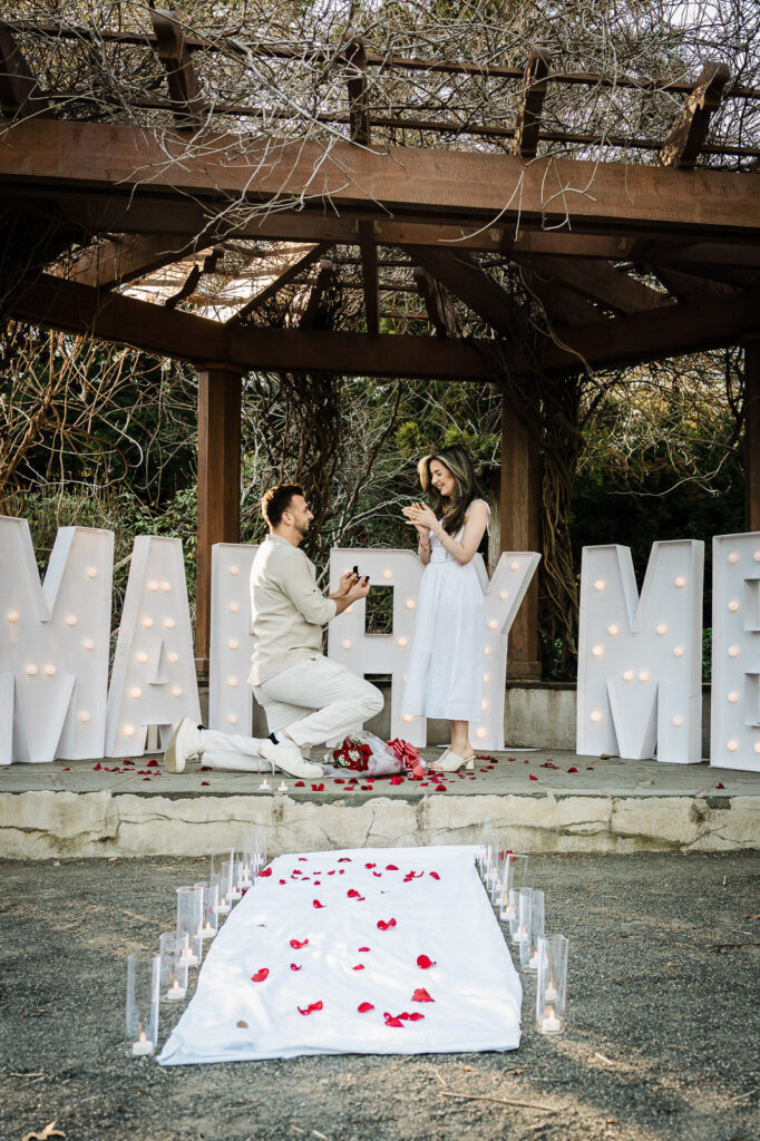 Man kneeling and presenting ring box during surprise proposal at Van Vleck House Gardens NJ