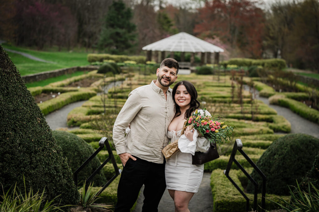 First kiss as an engaged couple in the Deep Cut Gardens parterre — Dryellen holding a spring bouquet, pavilion behind them 