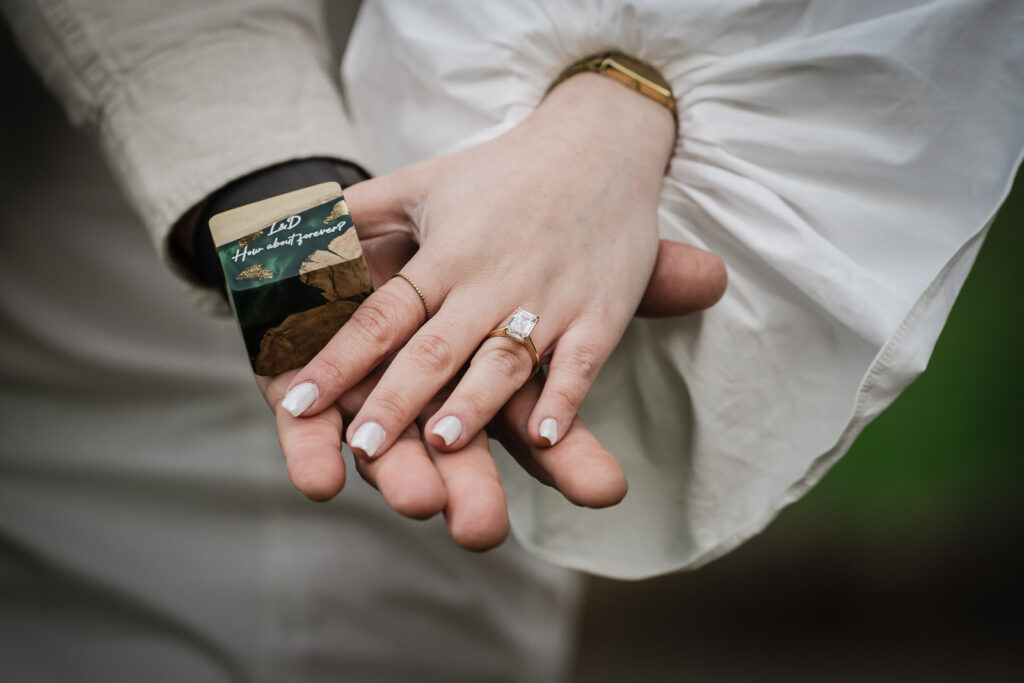 Emerald cut engagement ring on Dryellen's hand with custom proposal card reading L&D How about forever — Deep Cut Gardens NJ
