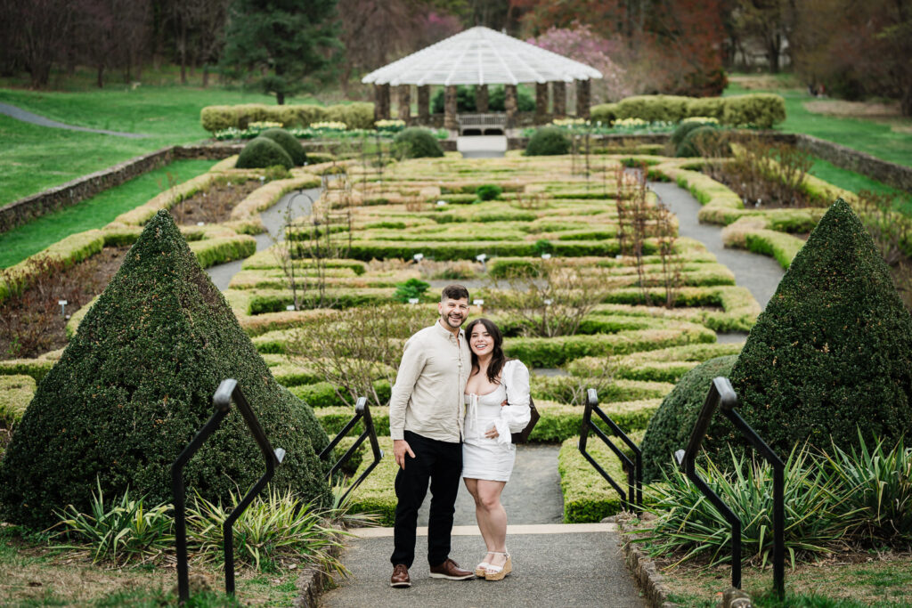 Luiz and Dryellen holding hands and smiling in the Deep Cut Gardens formal garden — stone pavilion visible in background