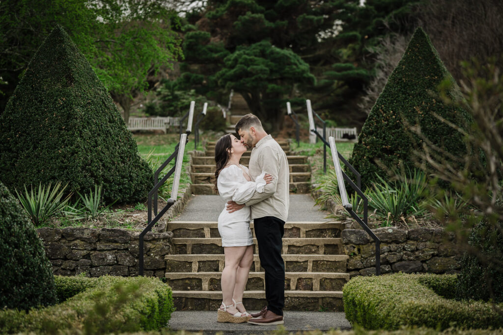 Luiz and Dryellen kissing at the stone staircase at Deep Cut Gardens NJ — topiary pyramids flanking them