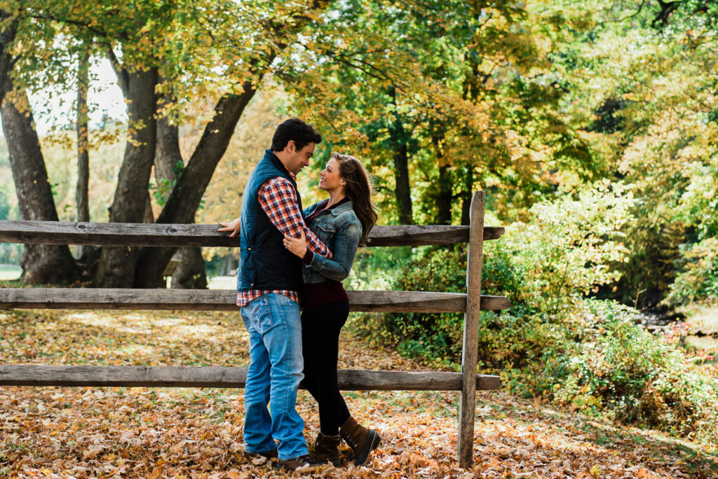 Engaged couple in fall layers leaning against rustic wooden fence at Natirar New Jersey in autumn