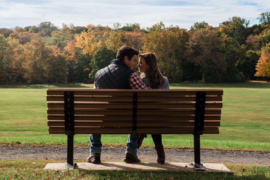 Engaged couple sitting on bench at Natirar with golden fall foliage filling the background