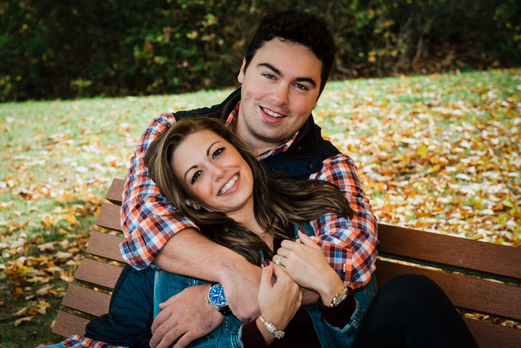 Engaged couple laughing and embracing on a bench surrounded by autumn leaves at Natirar NJ