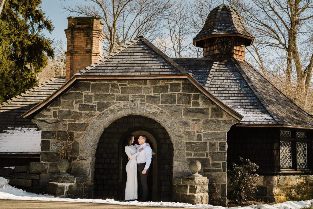 Couple embracing in the archway of the historic stone carriage house at New Jersey Botanical Garden Skylands Manor during a winter engagement session photographed by Alex Kaplan