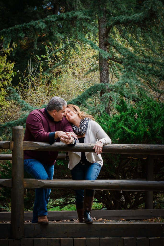 Couple leaning on wooden bridge railing holding hands during intimate engagement session at Overpeck County Park