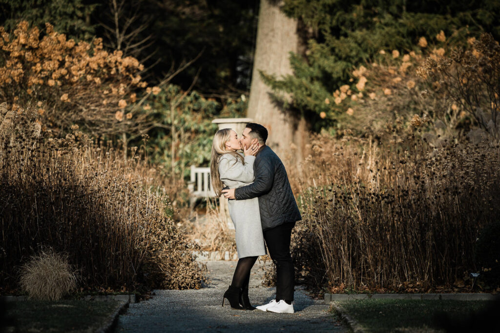 Couple sharing a romantic kiss along a garden path at Van Vleck House and Gardens in Montclair New Jersey during a winter proposal session photographed by Alex Kaplan