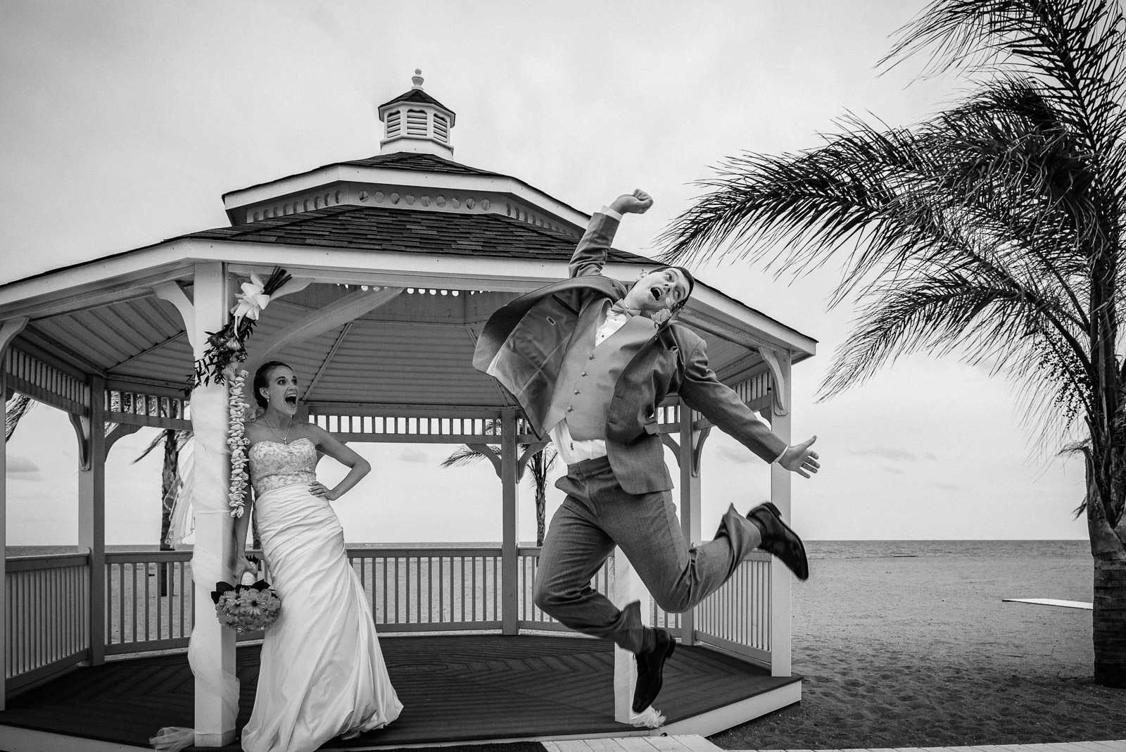 Unplanned wedding photo of a groom jumping for joy beside his laughing bride at a beachfront gazebo