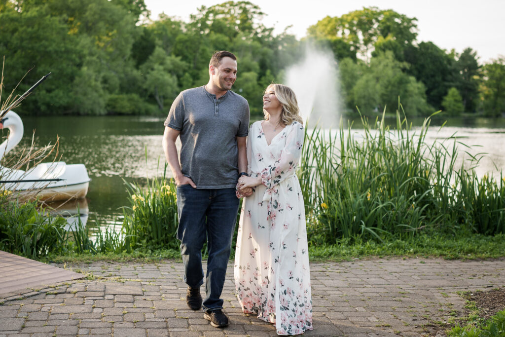 Couple walking hand in hand along the lake at Verona Park New Jersey with the swan boats and fountain visible behind them photographed by Alex Kaplan