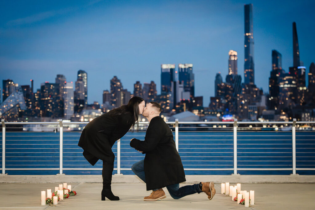 Couple sharing a first kiss after a candle-lit proposal on a Weehawken New Jersey pier at dusk with the full Manhattan skyline glowing behind them photographed by Alex Kaplan