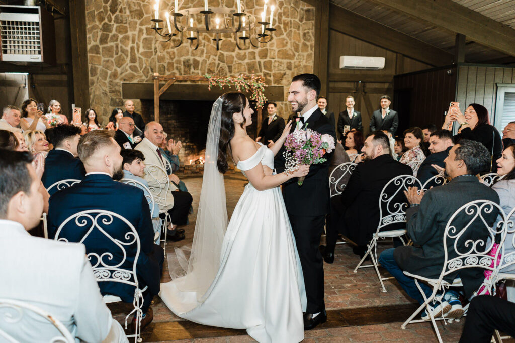 Kiara reaching up to touch Otari's chest as they share their first
joyful moment together at the Waterloo Village wedding altar surrounded
by guests and the historic stone fireplace