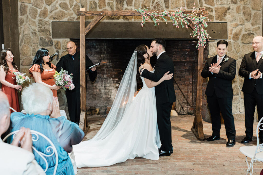 Kiara and Otari's first kiss as a married couple at the stone fireplace
altar at Waterloo Village wedding in New Jersey with guests applauding
on both sides