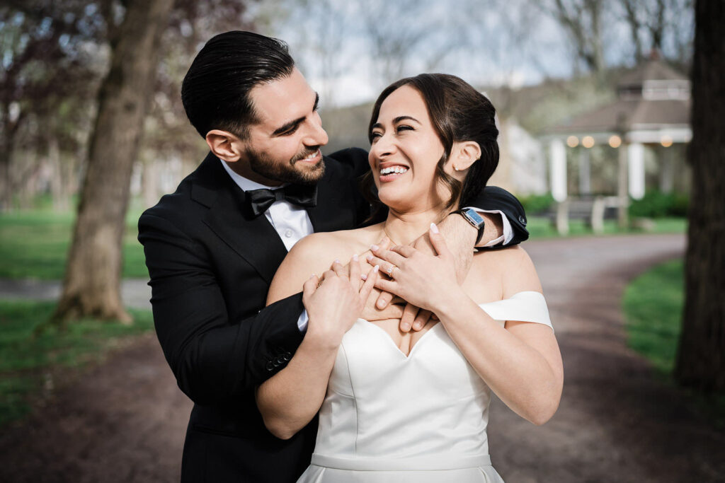 Groom holding bride from behind as they both laugh during outdoor portraits at Waterloo Village New Jersey with gazebo in background