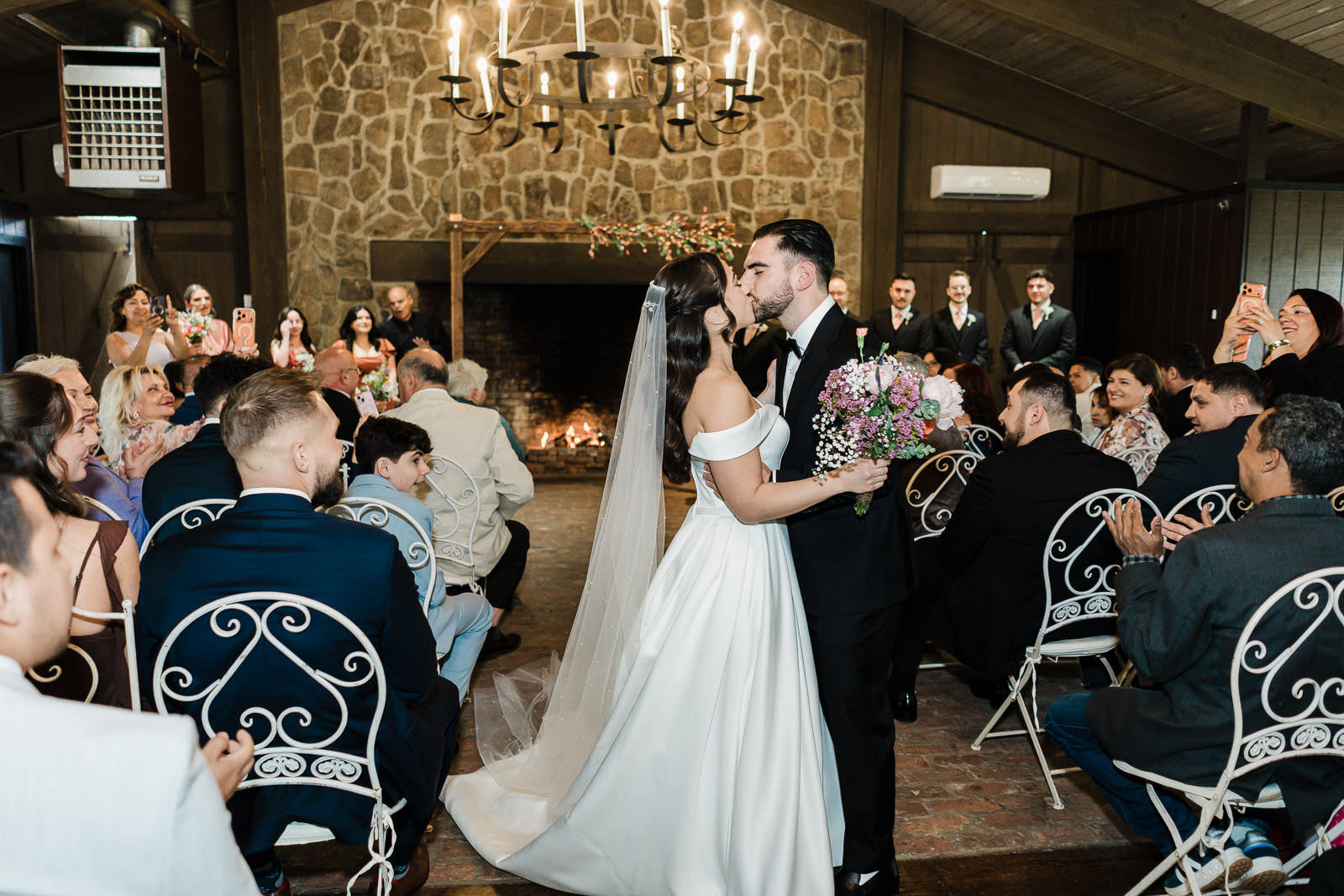 Wide view of Kiara and Otari's first kiss during their Waterloo Village wedding ceremony with guests celebrating in the rustic stone ceremony room in New Jersey