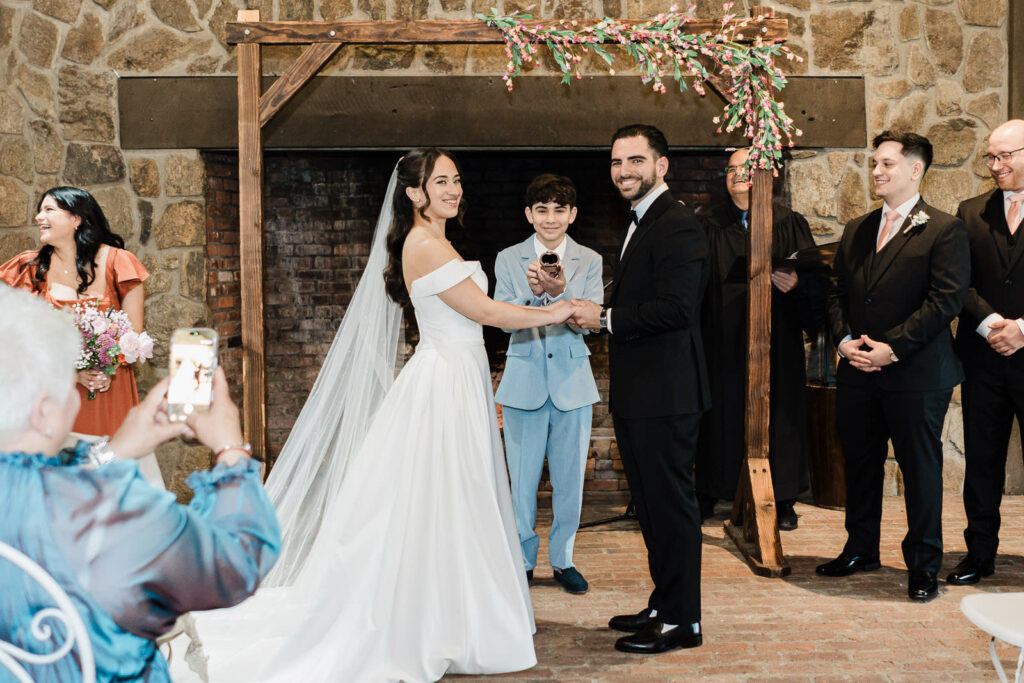 Ring bearer presenting the ring box to Kiara and Otari during their
ring exchange at the Waterloo Village wedding altar in front of the
stone fireplace and floral wooden arch