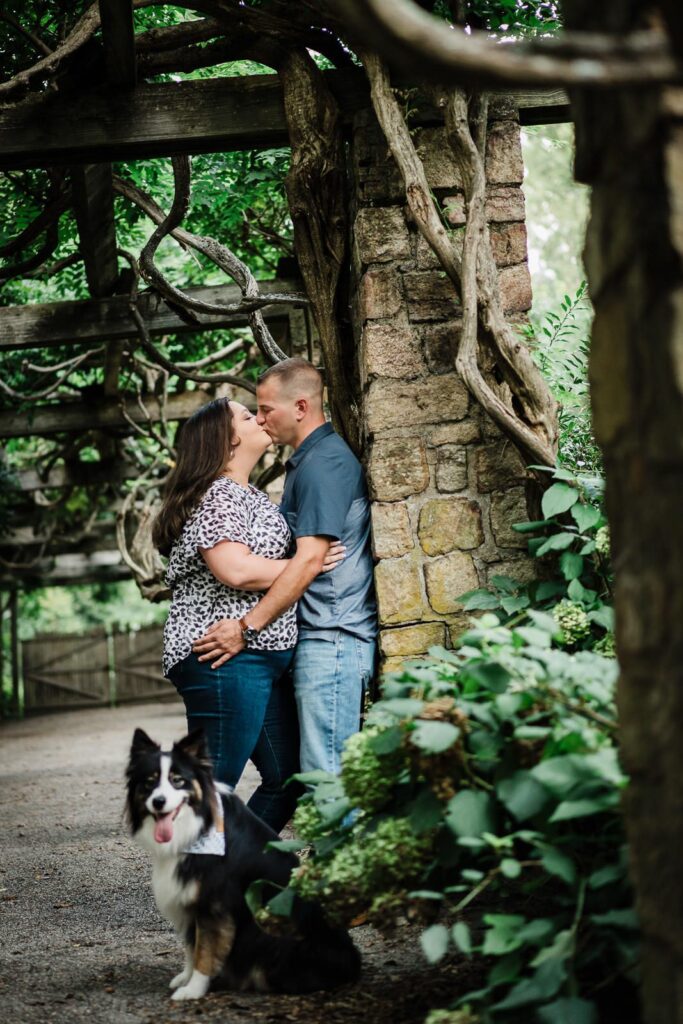 Couple kissing under the ivy-covered pergola with their dog at Crossed Keys Estate NJ