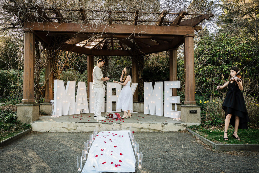 Marry Me marquee letters and petal aisle set up under a garden pergola for a proposal.