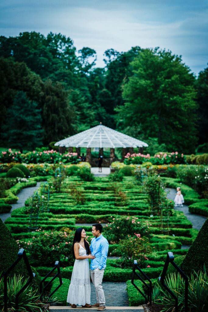 Engaged couple at Deep Cut Gardens with other park visitors in the garden behind them