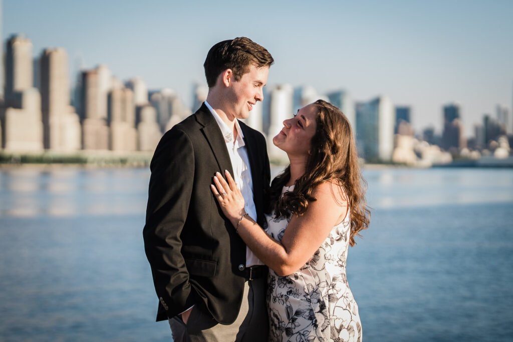 Newly engaged couple smiling at each other with Manhattan skyline and river behind