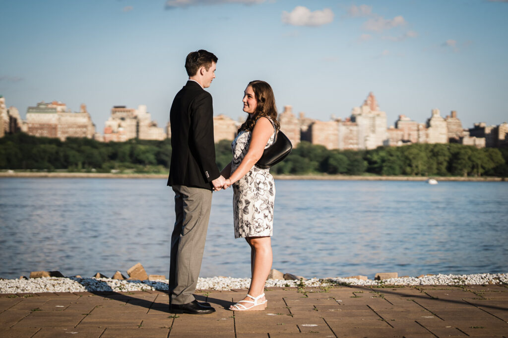 Engaged couple holding hands facing each other on waterfront walkway with Manhattan skyline