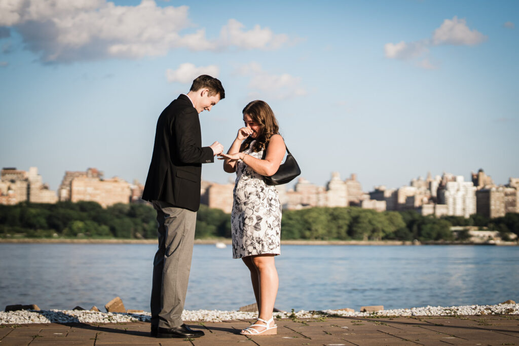 Man placing engagement ring on partner's finger on waterfront walkway after proposal