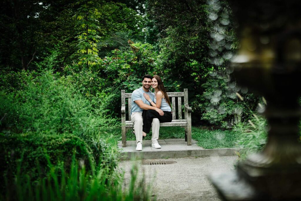 Engaged couple seated on a garden bench framed by foliage at Van Vleck Gardens