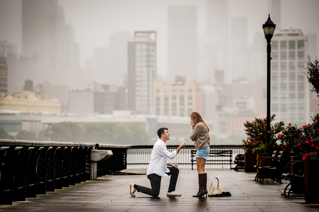 Man proposing on one knee as partner reacts on a rainy Hoboken pier in New Jersey