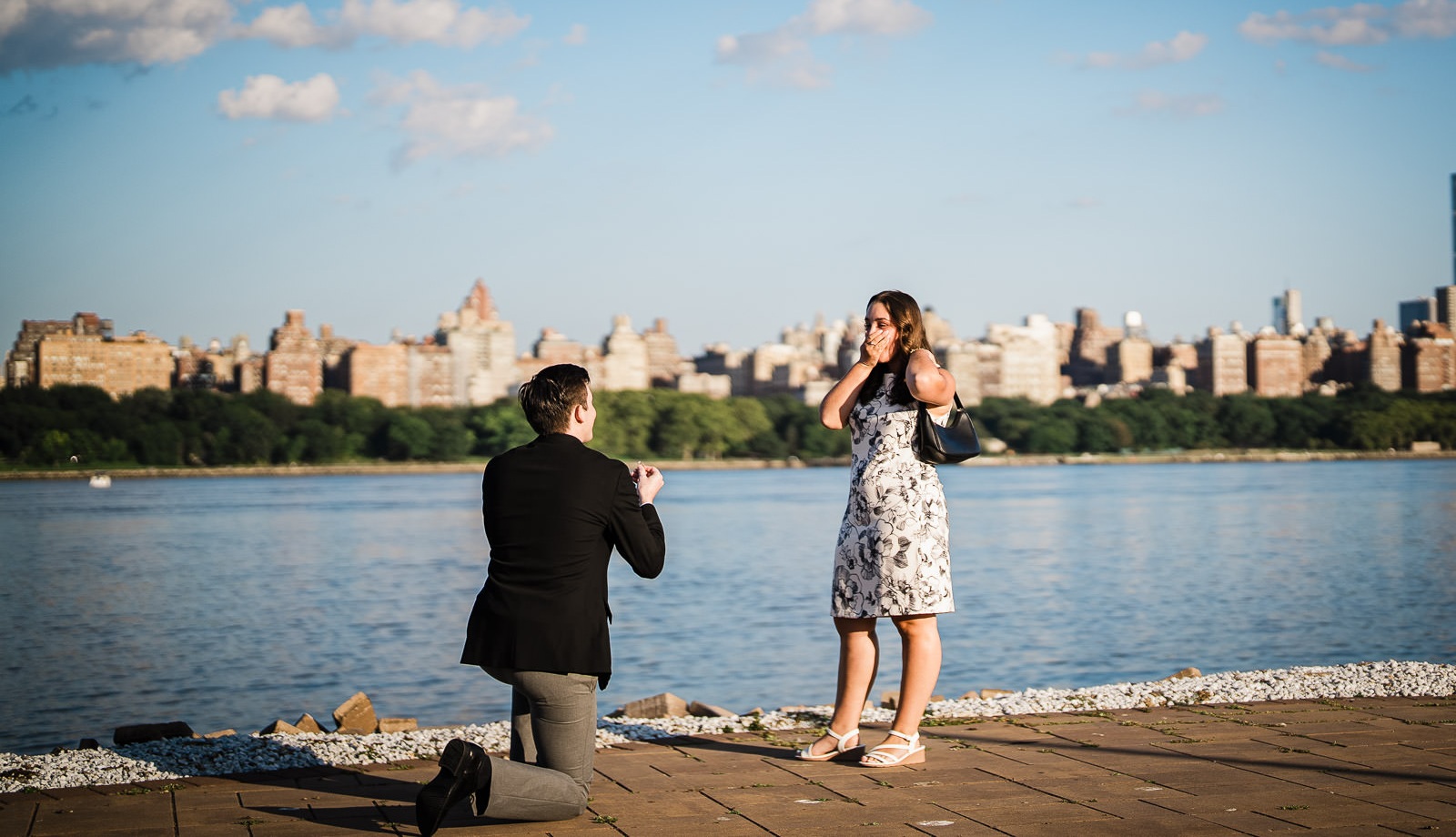 Man proposing on one knee to partner on waterfront walkway with Manhattan skyline behind