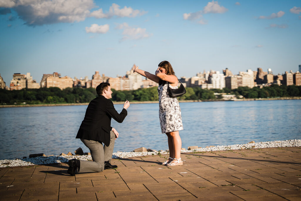 Woman reacting with one hand to her face as partner proposes on waterfront walkway