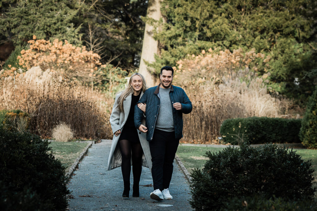 Couple in coordinated coats walking a garden path before a proposal in Montclair New Jersey