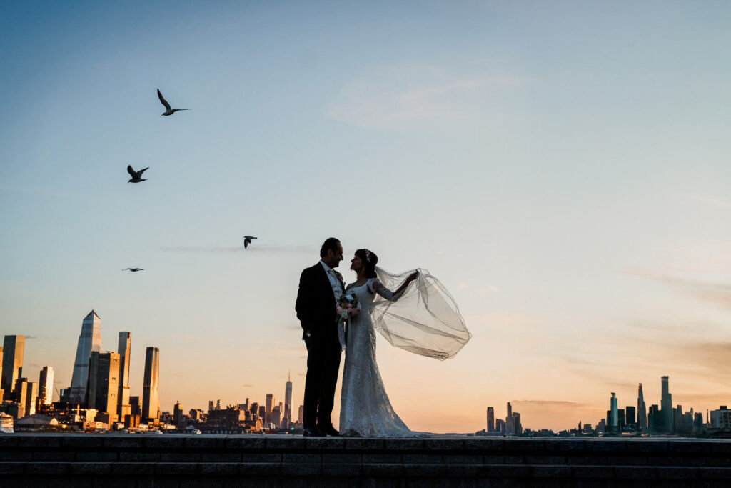 Bride and groom silhouetted against sunset skyline with birds flying overhead

